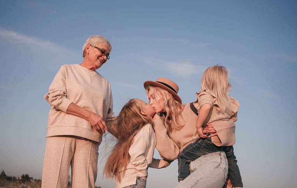 Females in family, grandma, daughter, and two granddaughters, all wearing matching beige and jeans with light hair standing in front of blue sky with grandma gazing down to daughter kissing granddaughter