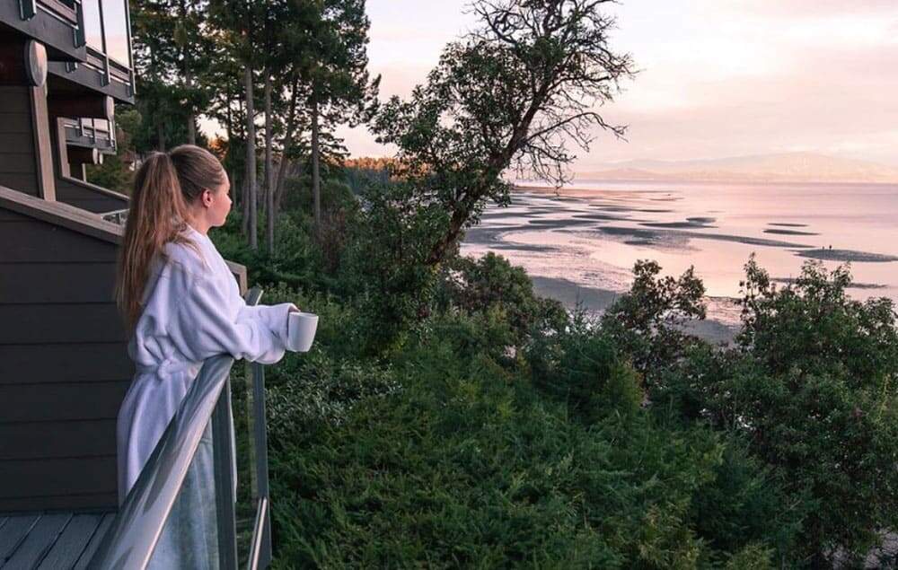 Woman with tied up long brown hair looking out of balcony with mug in hand and wearing white robe looking out to forest and beaches